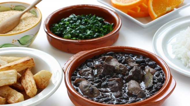 Imagem de um prato de feijoada com outros pratos de acompanhamentos como arroz e fatias de laranja